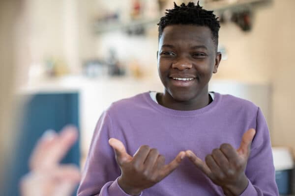 A young man wearing a purple shirt speaking sign language and smiling.